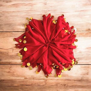 Puede incluir: Un pañuelo rojo vibrante, ingeniosamente dispuesto, adornado con lentejuelas doradas y pequeñas cuentas blancas. El pañuelo está drapeado en un patrón circular, creando un diseño floral.
