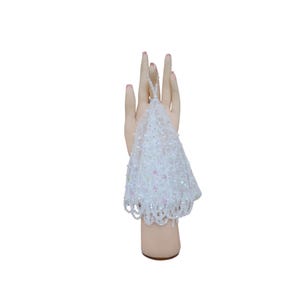 May include: A white beaded accessory, possibly a glove or wristlet, is shown on a mannequin hand. The item has detailed beadwork and a scalloped edge, with hints of pink. The background is plain white.