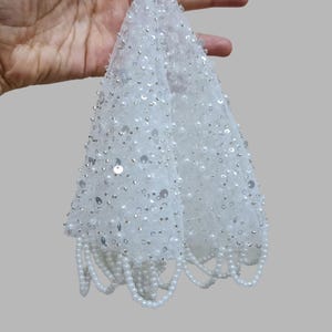 May include: A white, beaded evening bag with a delicate net overlay. The bag is decorated with sequins and small pearls, with a fringe of pearls along the bottom edge. The bag is held up by a hand.