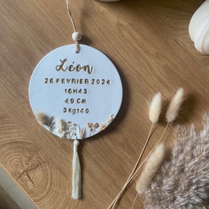 Puede incluir: Adorno redondo de cerámica blanca con letras doradas. El adorno dice "Léon" e incluye la fecha "26 FEVRIER 2024", la hora "16H43", la longitud "49 CM" y el peso "3KG140". El adorno está decorado con flores secas y tiene un borla blanca.