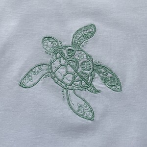 May include: A green embroidered sea turtle design on a white fabric background.