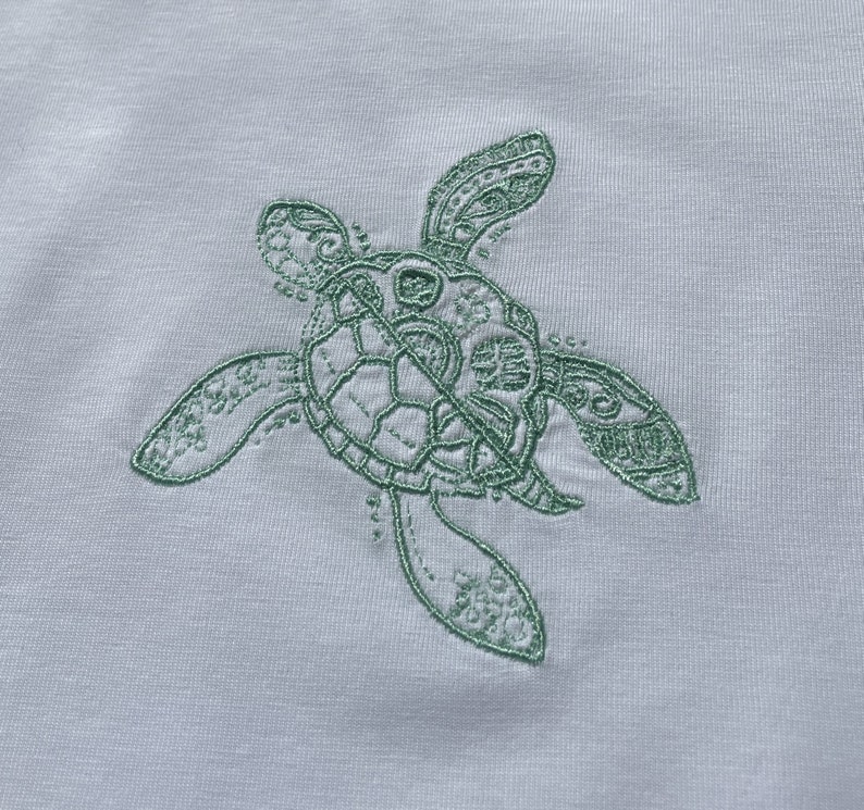 May include: A green embroidered sea turtle design on a white fabric background.