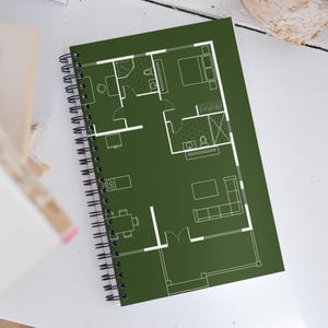 以下が含まれることがあります： 白い建築平面図が描かれた緑色の表紙の、スパイラル綴じノート。平面図には、部屋、家具、備品の輪郭が含まれています。ノートは開かれており、詳細なデザインが明らかになっています。