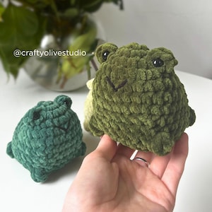 May include: Two handmade crochet frog plushies in shades of green. The larger frog is held in a hand, showcasing its textured, plush design. The smaller frog sits beside it, both with stitched facial features.