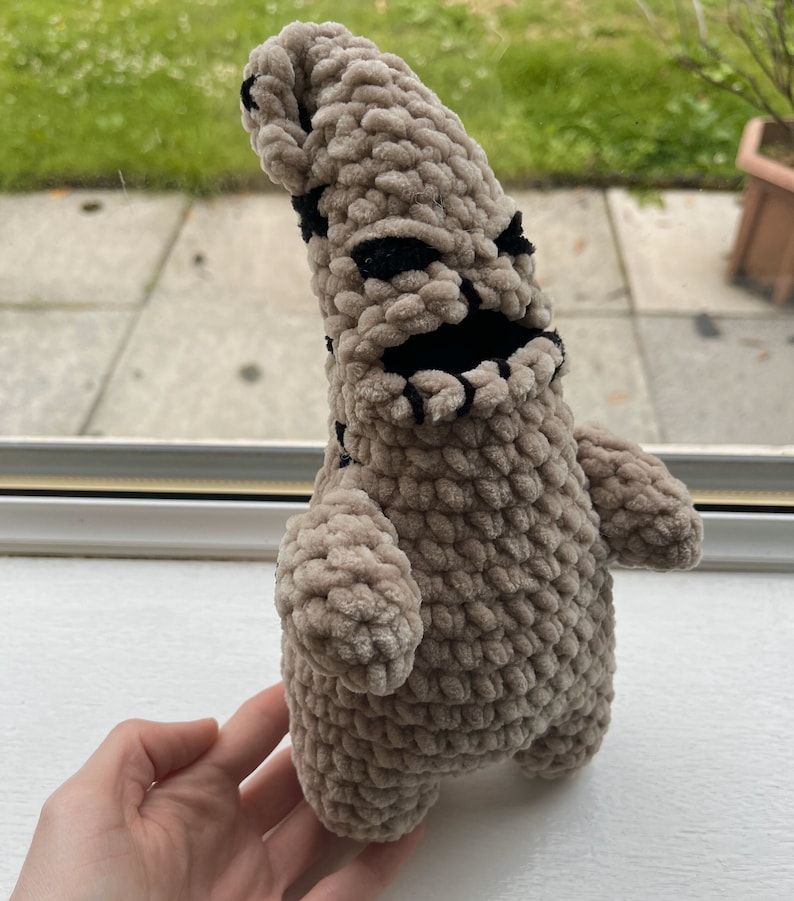 May include: A crocheted plush toy resembling a character from the movie "The Nightmare Before Christmas". The toy is brown with black facial features and is standing on a white surface.