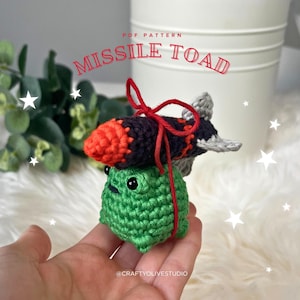 May include: A handmade crochet toy featuring a green frog with a missile on its back. The missile is black and orange. The text "MISSILE TOAD" is displayed in red. The toy is held in a hand.
