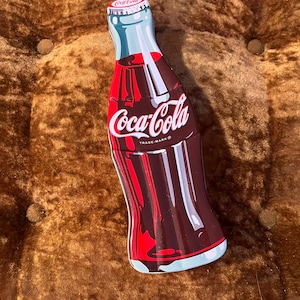 May include: A bottle-shaped metal tin featuring a dark brown and red graphic of a Coca-Cola bottle. The design includes the white Coca-Cola logo in script across the centre and a smaller logo on the bottle cap, with light blue highlights.