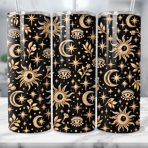 May include: A cylindrical tumbler with a black background featuring a repeating pattern of gold-colored suns, crescent moons, stylized eyes, and stars. The design includes small floral sprigs and a clear straw inserted at the top.