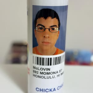 May include: White tumbler featuring a portrait of a man with glasses and the name "McLOVIN" printed on it. The tumbler also includes a barcode and the address "892 MOMONA ST HONOLULU, HI". The words "CHICKA CHICKA" are printed at the bottom.