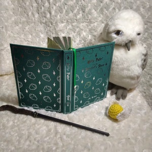 May include: A green hardcover book with a silver pattern of lightning bolts and gemstones. The book is titled "Harry Potter and the Sorcerer's Stone" and has the author's name, J.K. Rowling, on the cover. The book is open to reveal the title page, which reads "Harry Potter Year One". A brown wooden wand and a white stuffed owl are also in the image.