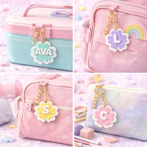 May include: A collection of personalized flower-shaped keychains with gold-colored chains. Each keychain features a different initial or name, with pastel colors like pink, blue, yellow, and purple. The keychains are attached to a lunchbox, backpack, and pencil case.