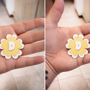 May include: A layered flower-shaped pendant with a yellow center, white petals, and a white letter "D" in the center. The pendant has a small hole at the top for hanging. The pendant is held in a person's hand.