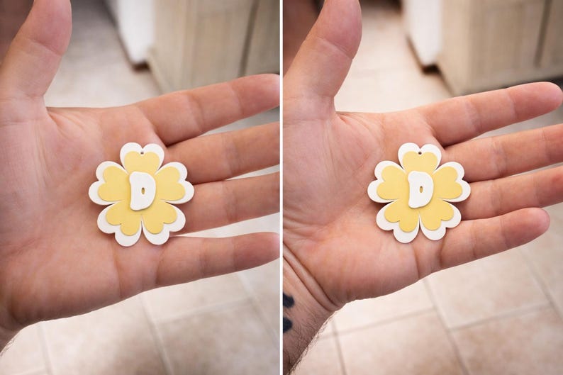 May include: A layered flower-shaped pendant with a yellow center, white petals, and a white letter "D" in the center. The pendant has a small hole at the top for hanging. The pendant is held in a person's hand.