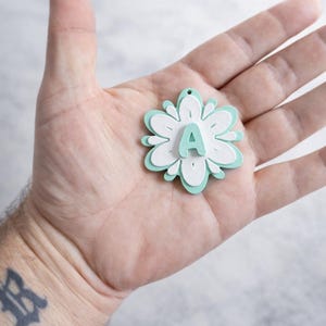 May include: A layered flower-shaped pendant with the letter "A" in the center. The pendant features a light blue and white color scheme. The pendant is held in a hand.