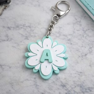 May include: A flower-shaped keychain with a silver chain and clasp. The keychain is white and light blue, with the letter "A" in the center. The keychain is on a marble surface, with keys and a notebook in the background.
