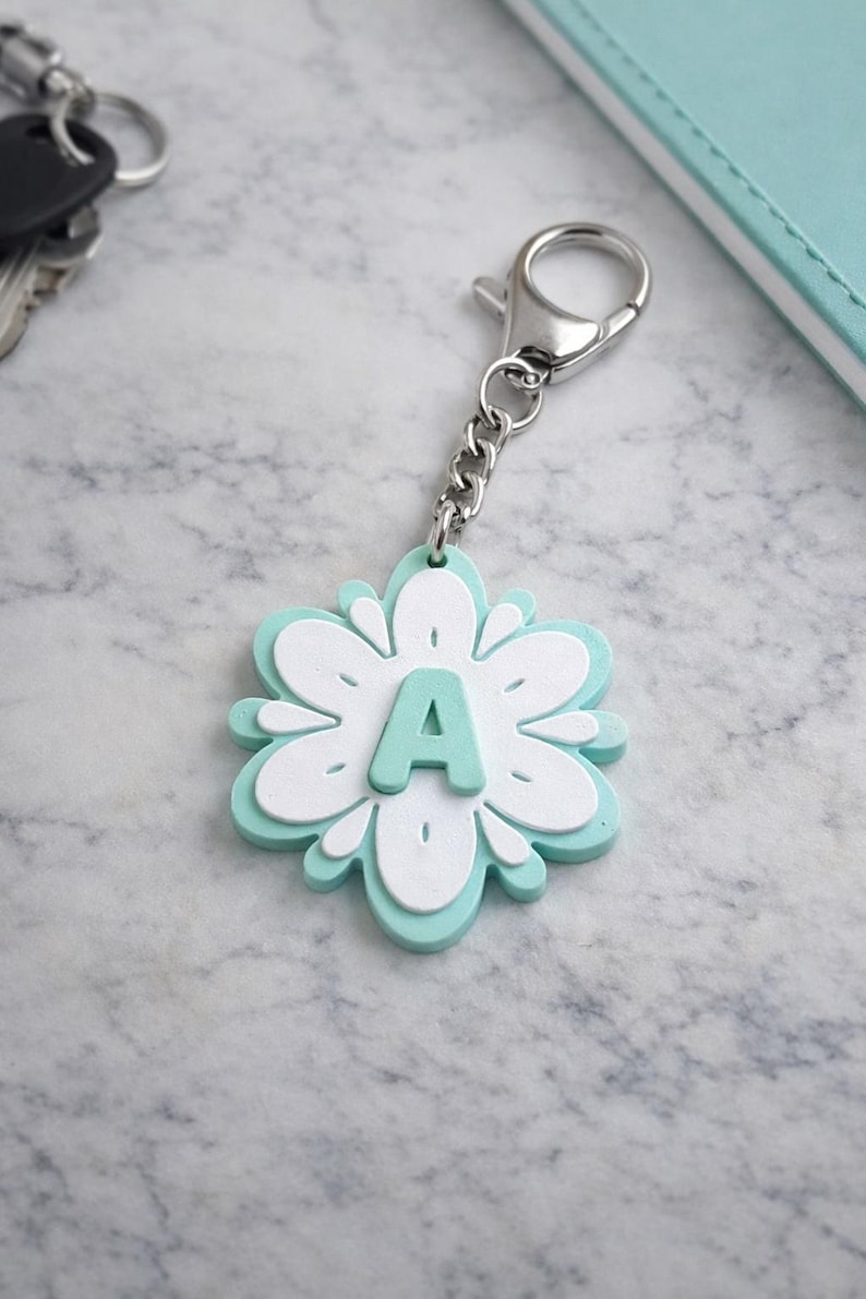 May include: A flower-shaped keychain with a silver chain and clasp. The keychain is white and light blue, with the letter "A" in the center. The keychain is on a marble surface, with keys and a notebook in the background.