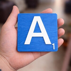 A hand holds a blue Scrabble tile with a white letter "A" and the number "1" in the bottom right corner.