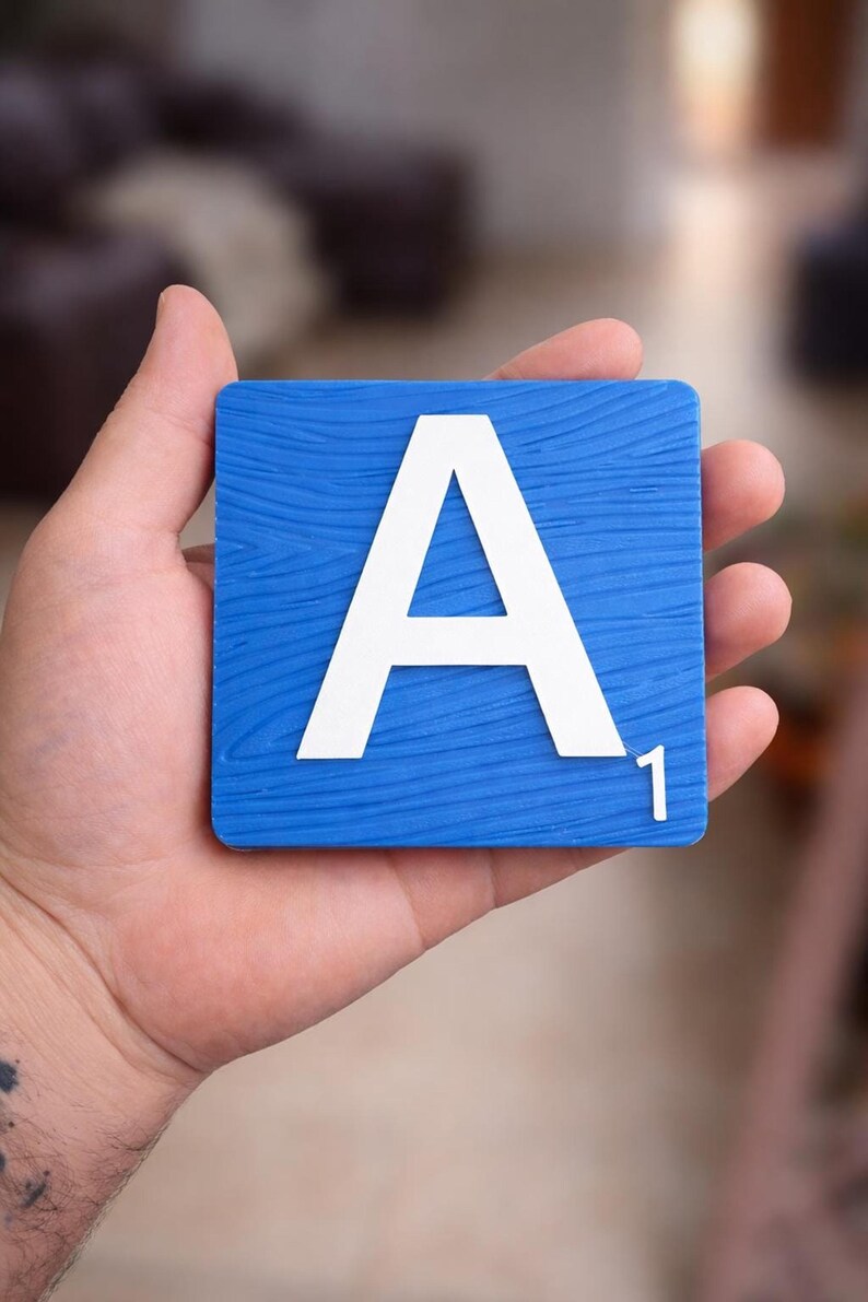 A hand holds a blue Scrabble tile with a white letter "A" and the number "1" in the bottom right corner.