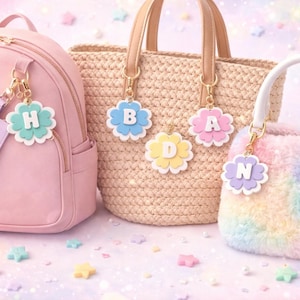 May include: A pink backpack, a woven tote bag, and a fluffy rainbow purse are displayed. Each bag has a flower-shaped charm with a letter on it. The charms are in various pastel colors, and the background is decorated with star-shaped confetti.