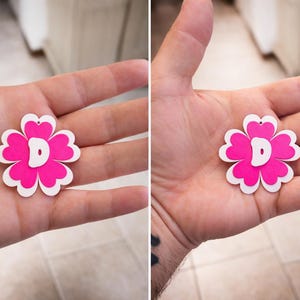 May include: A pink and white flower-shaped pendant with a white letter "D" in the center. The pendant is held in an open hand. The flower has a heart-shaped cutout at the top for hanging.