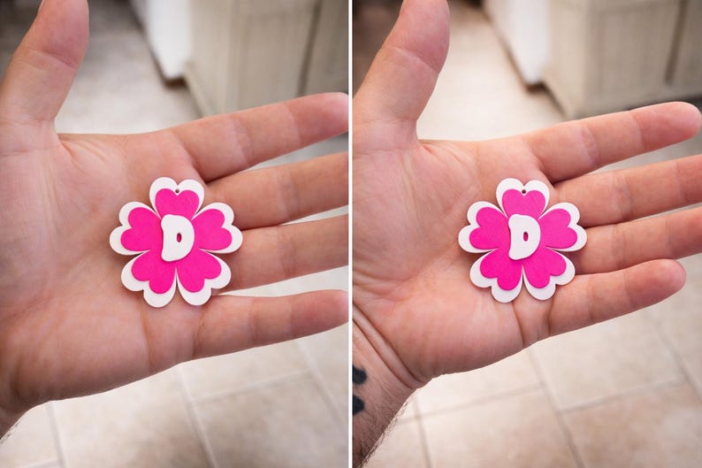 May include: A pink and white flower-shaped pendant with a white letter "D" in the center. The pendant is held in an open hand. The flower has a heart-shaped cutout at the top for hanging.