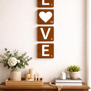 Four brown Scrabble tiles spell "LOVE" vertically on a white wall above a wooden console table with decor.