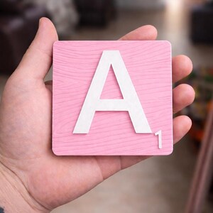 A hand holds a pink Scrabble tile with a white letter "A" and the number "1" in the bottom right corner.