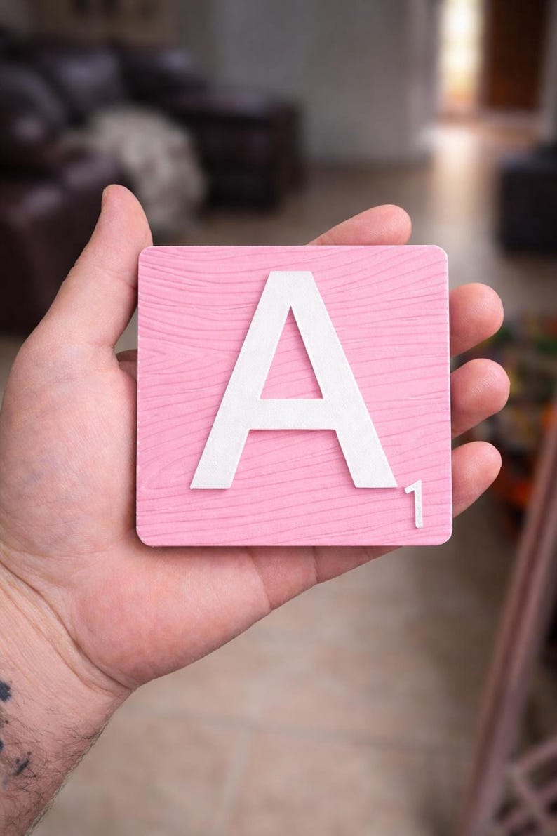 A hand holds a pink Scrabble tile with a white letter "A" and the number "1" in the bottom right corner.