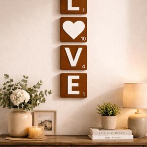 Four brown Scrabble tiles spell "LOVE" vertically on a white wall above a wooden console table with a lamp and other decor.