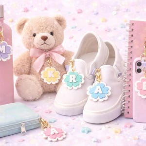May include: A collection of pastel-colored accessories, including a pink water bottle, a teddy bear, white sneakers, a phone, and a pencil case. Each item has a flower-shaped charm with a letter. The background is decorated with star-shaped confetti.