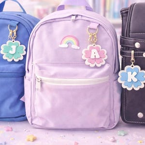 May include: Three backpacks in blue, lavender, and black. Each backpack has a flower-shaped charm with a letter: J, A, and K. The lavender backpack has a rainbow sticker. The charms are white, with pastel colors.