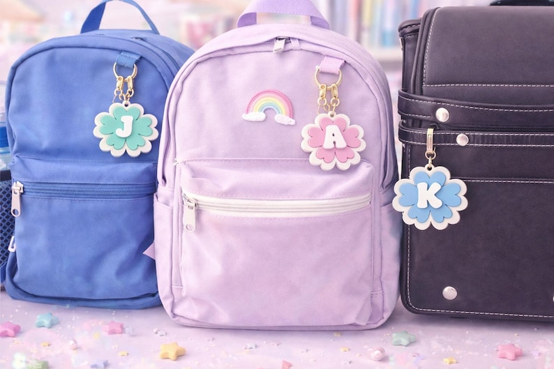 May include: Three backpacks in blue, lavender, and black. Each backpack has a flower-shaped charm with a letter: J, A, and K. The lavender backpack has a rainbow sticker. The charms are white, with pastel colors.