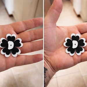 May include: A black and white flower-shaped charm with a letter "D" in the center. The charm is held in a hand, showcasing its design. The flower has a layered design with a white outline and a black center.