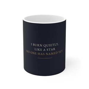 May include: A matte black ceramic mug with a white interior. The front features the gold-colored text: I BURN QUIETLY, LIKE A STAR NO ONE HAS NAMED YET. A thin horizontal gold line is positioned directly underneath the printed text.