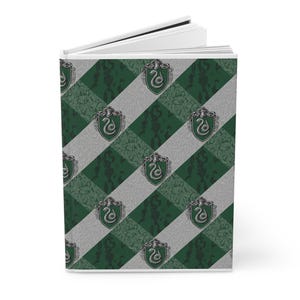 Puede incluir: Un cuaderno de tapa dura con un patrón diagonal verde y gris. El patrón incluye el escudo de Slytherin, una serpiente dentro de un escudo. El cuaderno está abierto, revelando páginas blancas.