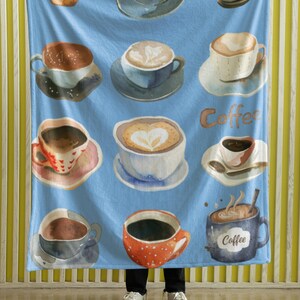 Pode incluir: Uma manta de lã azul com um padrão de aquarela de xícaras de café. As xícaras estão cheias de diferentes tipos de café, incluindo cappuccino, latte e espresso. A manta apresenta a palavra "Coffee" em uma fonte estilizada.