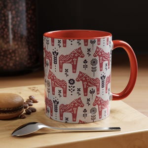 May include: A red and white ceramic mug with a pattern of red and black Dala horses and floral designs. The mug has a red interior and a red handle.