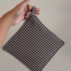 May include: A square-shaped potholder featuring a classic black and white gingham check pattern. The fabric item is finished with stitched edges and includes a small matching fabric loop at the top corner for convenient hanging and storage.
