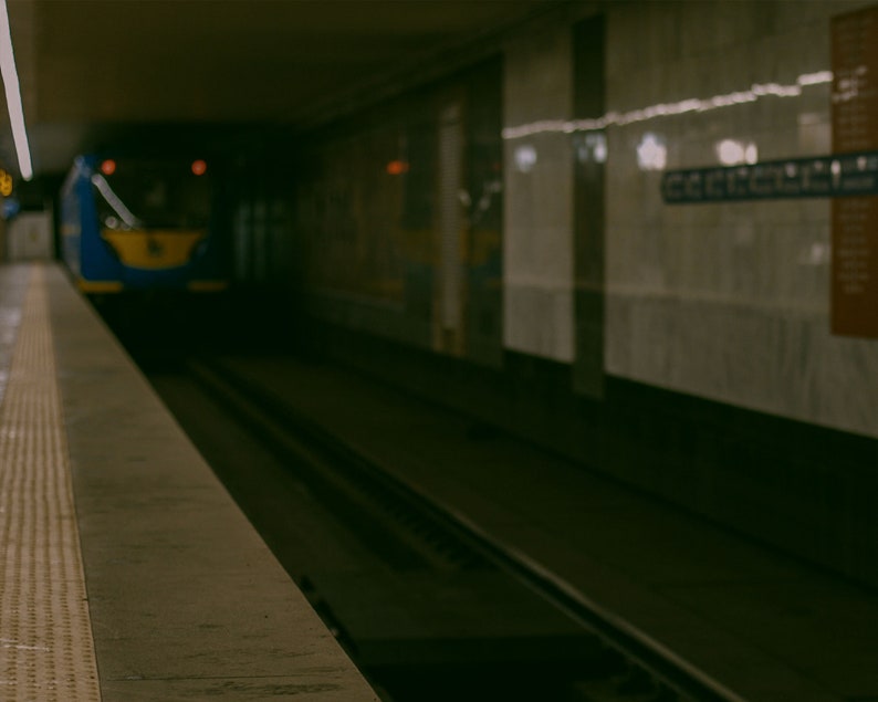 May include: A blue and yellow subway train approaches a station platform. The image shows the tracks and platform edge in the foreground, with a sign on the wall. The scene is dimly lit, suggesting an underground environment.