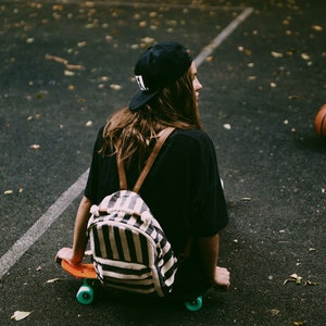 May include: A person wearing a black cap and shirt, sitting on an orange skateboard with teal wheels. A striped backpack with brown straps is on the person's back. A basketball is in the background.