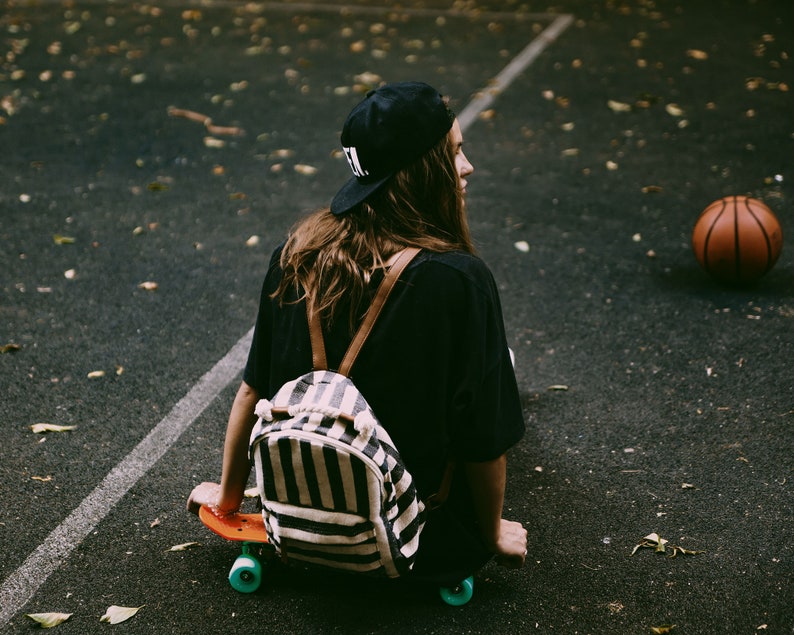 May include: A person wearing a black cap and shirt, sitting on an orange skateboard with teal wheels. A striped backpack with brown straps is on the person's back. A basketball is in the background.