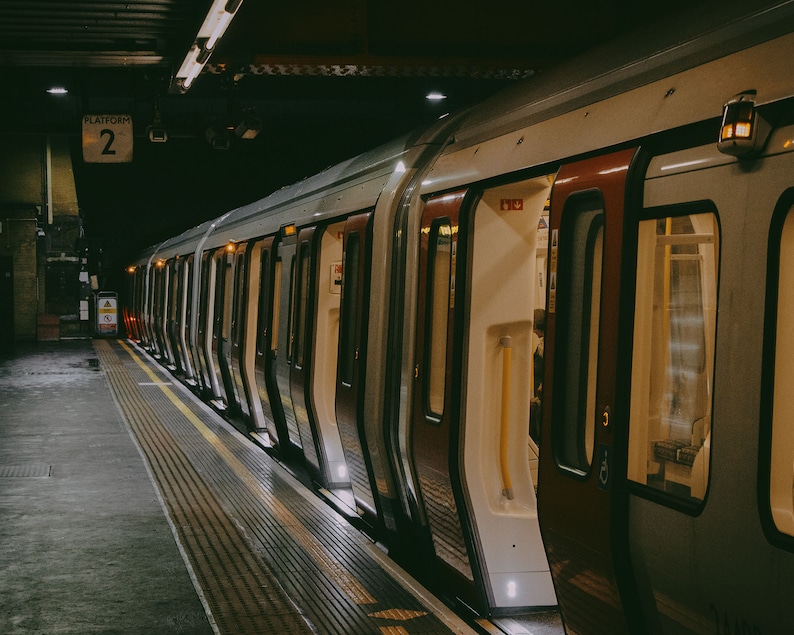 May include: A train carriage at a station platform with open doors. The train is silver and red, with interior lights visible. The platform has a yellow line and a sign that reads "Platform 2". The scene is dimly lit.