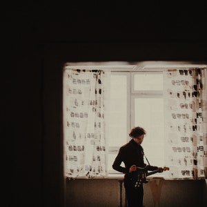 May include: A person playing an electric guitar in front of a window with patterned curtains. The person is wearing a dark suit. The room is dimly lit, with the window providing the primary light source.