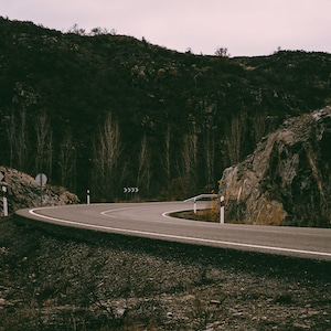 May include: A curved asphalt road winds through a mountainous landscape. The road is flanked by rocky cliffs and a forest of bare trees. Road signs are visible, and the sky is overcast.