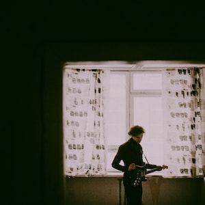 May include: A person playing an electric guitar in front of a window with patterned curtains. The image is in sepia tones, with the person in dark clothing and the guitar in black. The window provides a bright background.