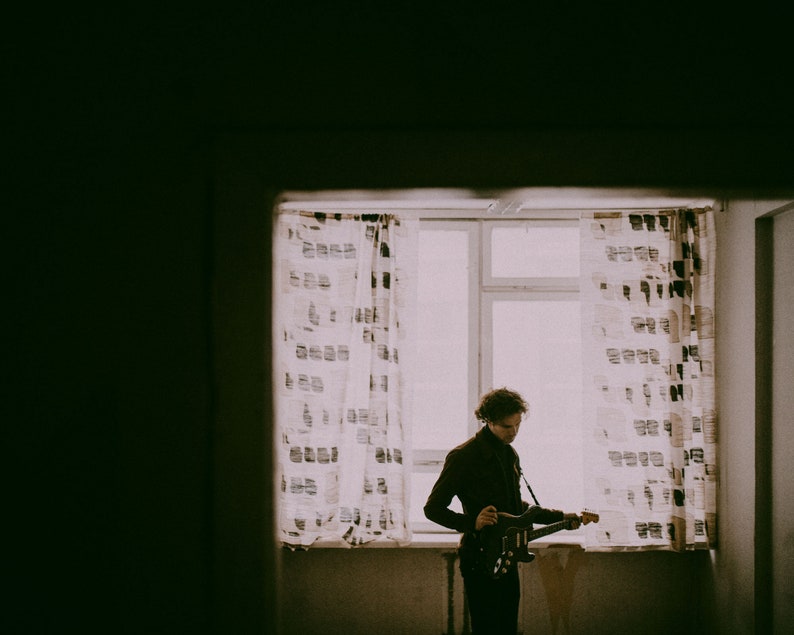 May include: A person playing an electric guitar in front of a window with patterned curtains. The image is in sepia tones, with the person in dark clothing and the guitar in black. The window provides a bright background.