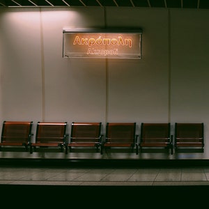 May include: A photograph of a subway station platform. Six wooden seats are arranged in a row. Above, a sign displays "Akropoli" in Greek and English. The lighting is dim, creating a sense of quiet and anticipation.