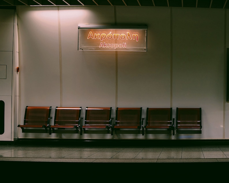 May include: A photograph of a subway station platform. Six wooden seats are arranged in a row. Above, a sign displays "Akropoli" in Greek and English. The lighting is dim, creating a sense of quiet and anticipation.