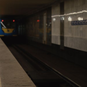 May include: A subway station platform with a train approaching in the tunnel. The train is blue and yellow. The platform has a textured edge. The walls are marble with a sign on the right.