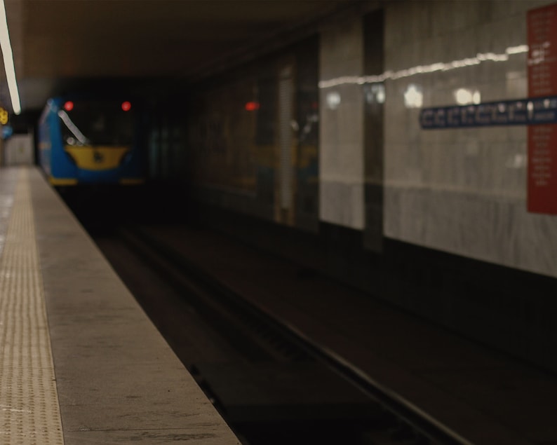 May include: A subway station platform with a train approaching in the tunnel. The train is blue and yellow. The platform has a textured edge. The walls are marble with a sign on the right.